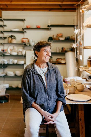 Happy middle aged female artisan looking away while resting after work in creative pottery studio