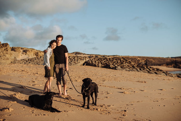 Millennial couple standing at beach with two black labradors