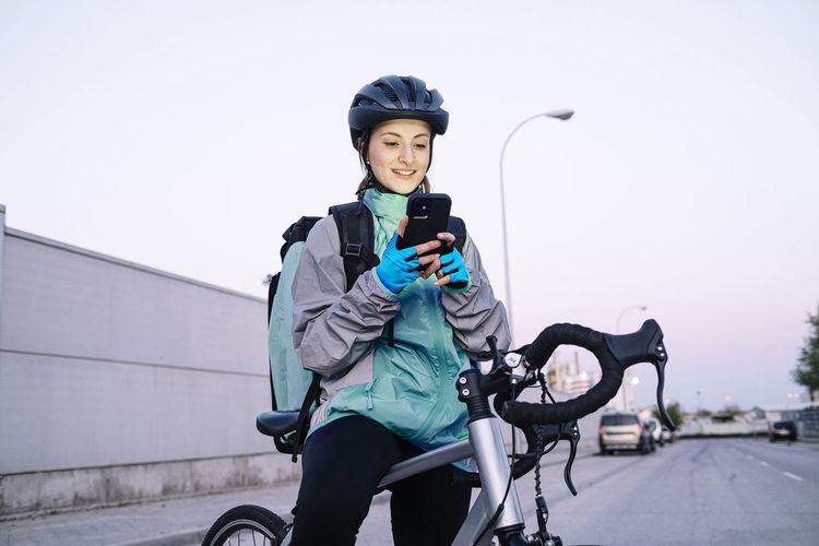 Young cheerful woman with thermal bag browsing smartphone with gps map while sitting on bicycle for delivering food on city street