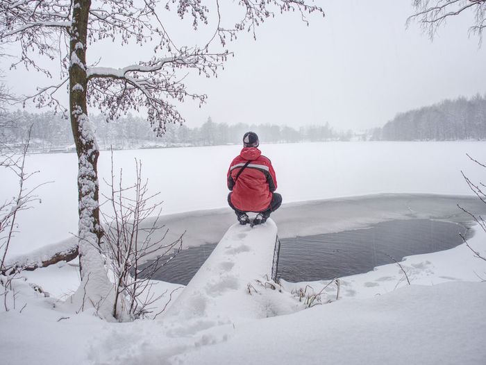 Man in red jacket stay at end of big pipe above dark water of unfrozen lake. winter under snow.