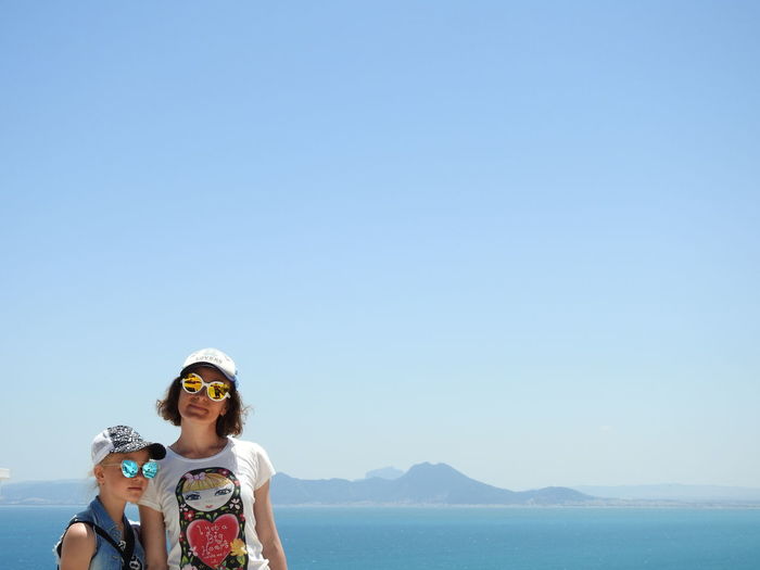 Mother and daughter wearing sunglasses standing against sea and clear blue sky during sunny day