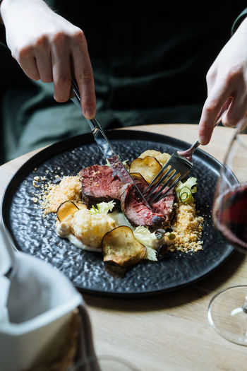 Crop from above of person with black plate with roast beef slices on creamy sauce with cauliflower and fried chips having dinner with glass of red wine