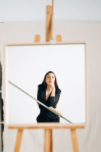 Young pensive female in black wear and earring looking at camera in mirror placed on easel
