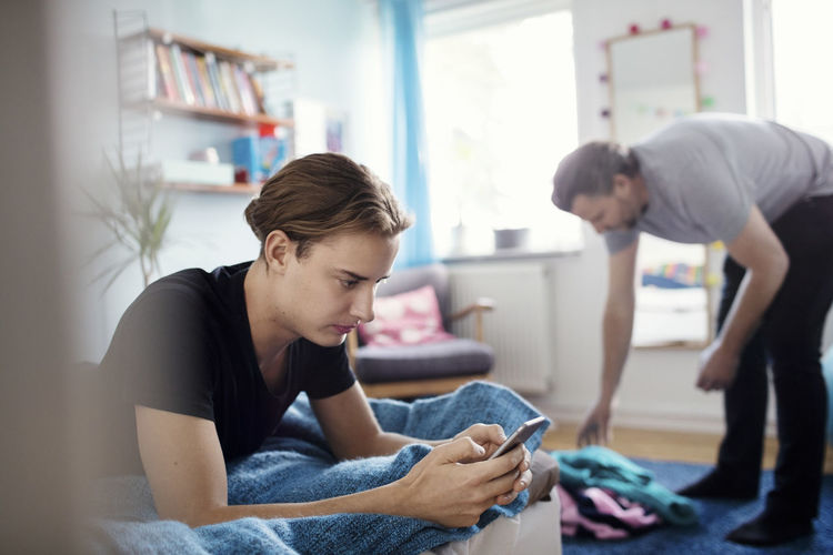 Man using smart phone while father picking clothes fallen on rug at home