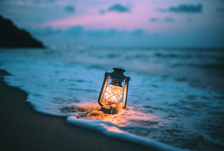 Illuminated lamp on beach against sky at sunset