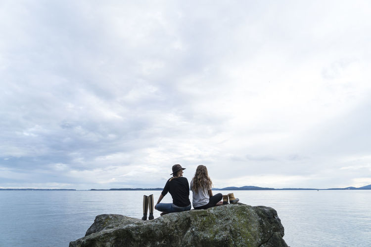 Two friends sit on a rock with their shoes beside them looking out over the water