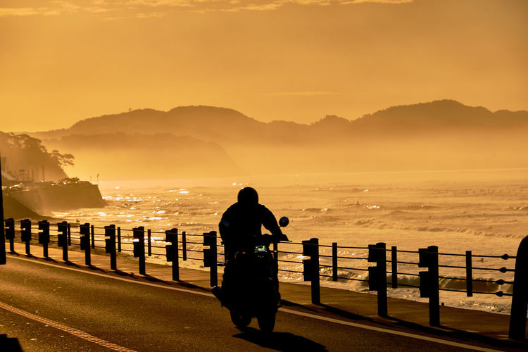 Silhouette person on motorcycle by railing against sky and sea during sunrise