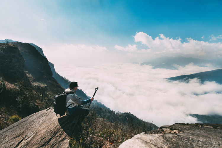 Man sitting on cliff against cloudy sky