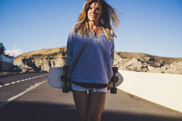 Portrait of smiling young woman with surfboard standing on road