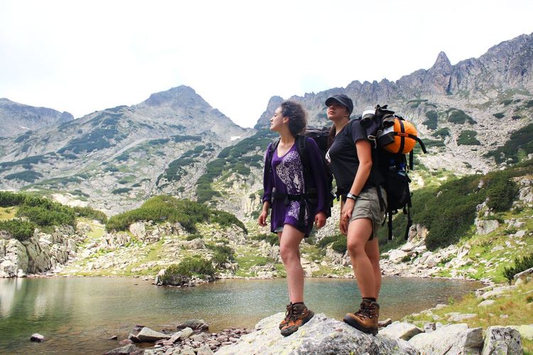 Low angle view of female backpack hikers standing at lakeshore against mountains
