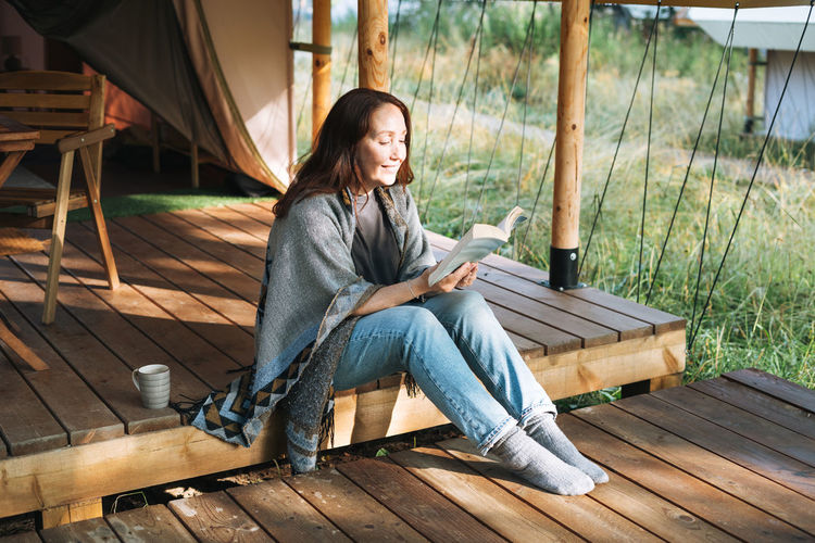 Young brunette woman in poncho reading book and relaxing in glamping in the woods