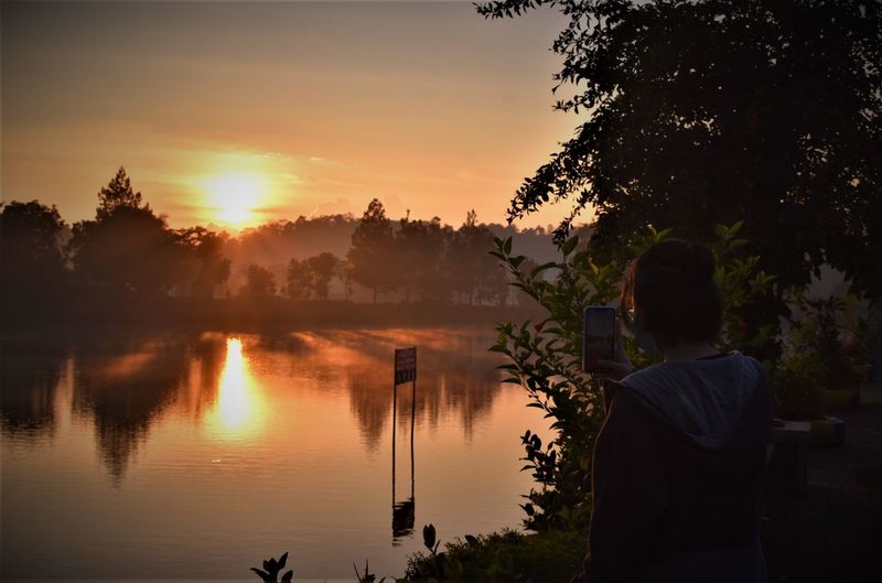 Rear view of silhouette man standing by lake against sky during sunset
