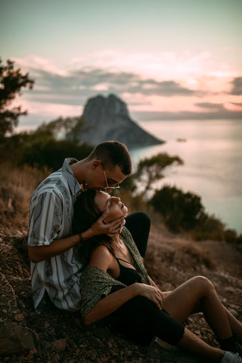 Midsection of couple sitting against sky during sunset