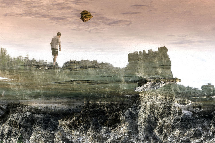 Reflection of man walking on rock in sea against sky