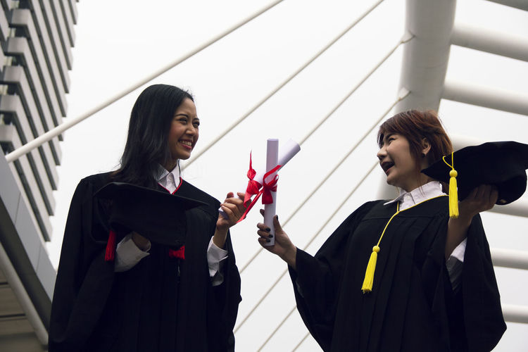 Smiling female friends wearing graduation gowns holding certificates