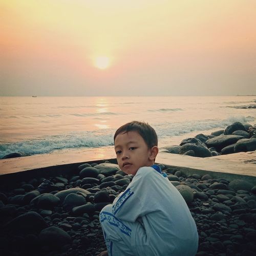 Cute boy crouching at beach against clear sky during sunset