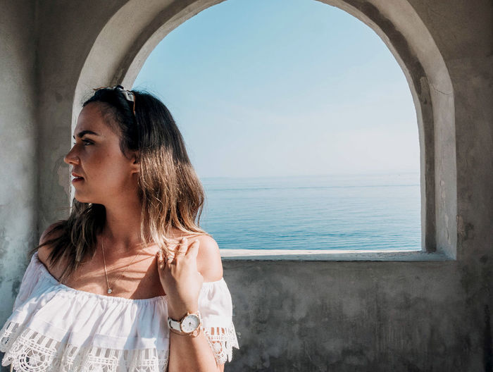 Portrait of young woman in front of arched window overlooking sea.