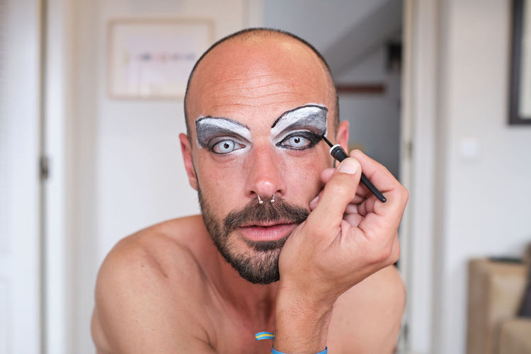 Adult shirtless bearded male transgender with eyebrow makeup and cosmetic lens applying black eyeliner