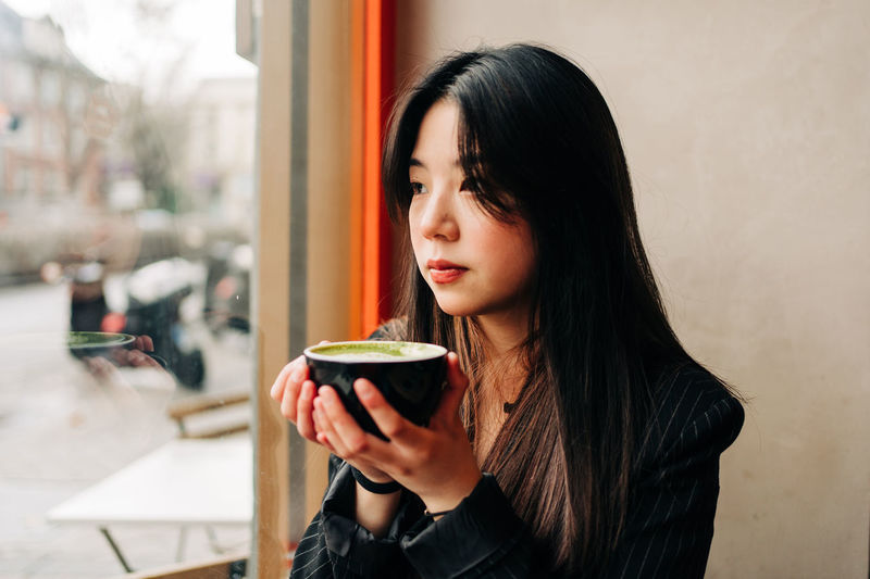 Long-haired brunette asian woman having a coffee on a coffee shop while is looking a cellphone through the window