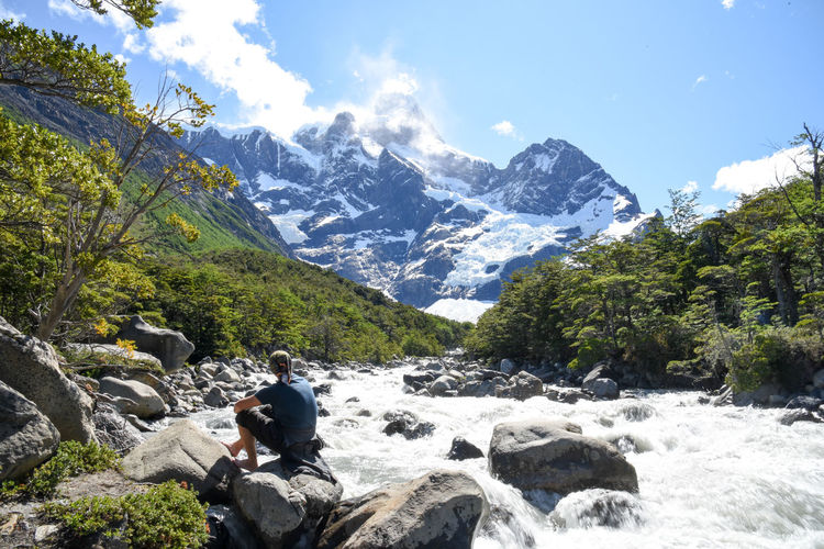 Man sitting on rock by river flowing against snowcapped mountains