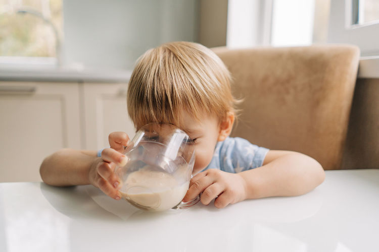 Little toddler girl drinks delicious fresh milk from a mug in the kitchen at home.
