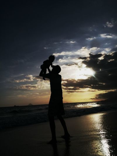 Silhouette of man holding up child against cloudy sky