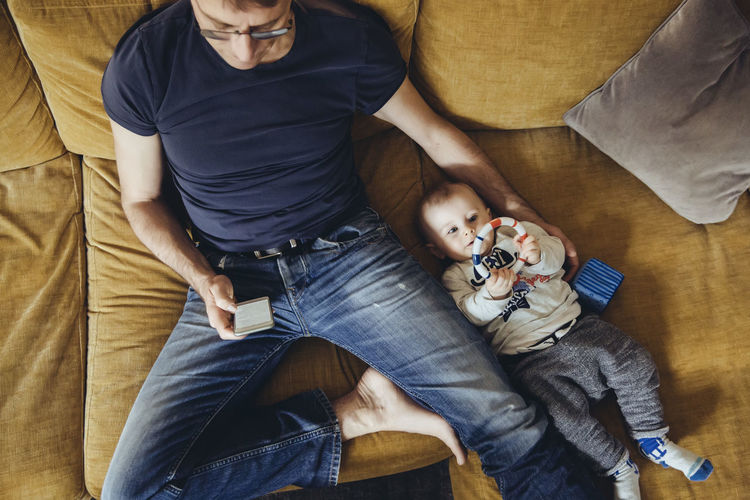 Baby boy lying on couch besides his father using smartphone