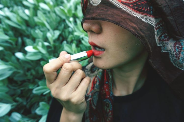 Close-up of woman applying red lipstick against plants