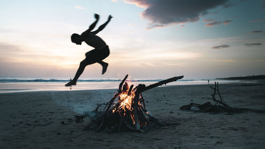 Full length of man jumping over bonfire at beach against sky during sunset
