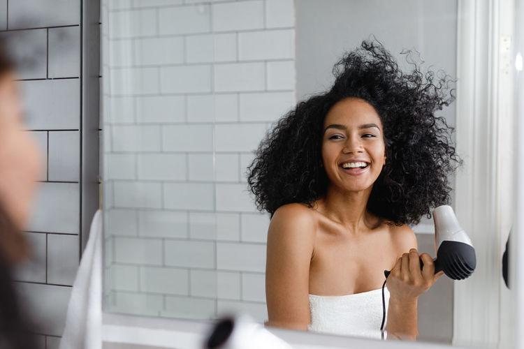 Smiling young woman drying hair reflecting on mirror