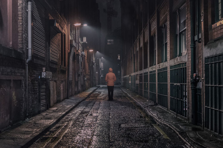 Rear view of mature woman walking on alley amidst buildings in city at night