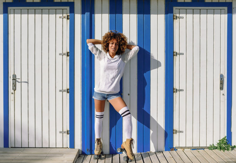 Young black woman on roller skates near a beach hut. Girl with afro hairstyle rollerblading on sunny promenade. Afro Adult Adults Only Afrohair Beach Huts Beautiful Woman Curly Hair Day Front View Full Length Hut Looking At Camera One Person One Woman Only One Young Woman Only Only Women Outdoors People Portrait Roller Skating Rollerblading Rollerskating Skates Young Adult Young Women