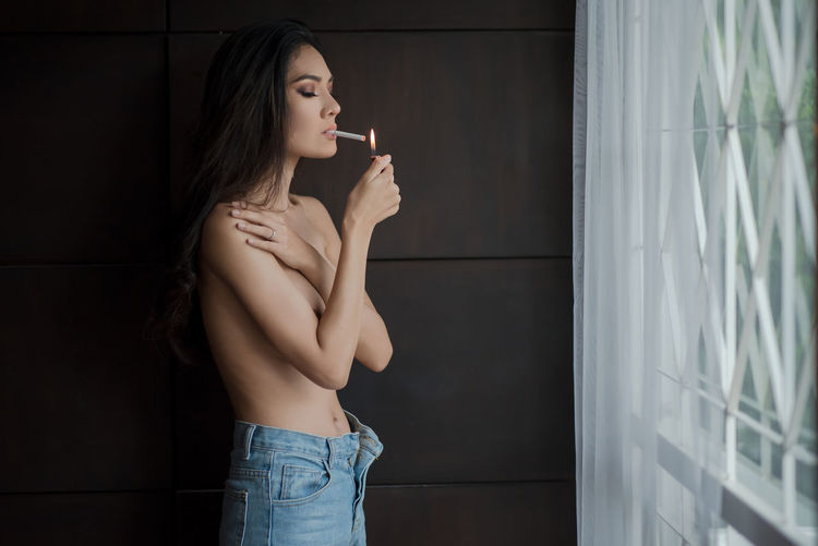 Side view of shirtless young woman lighting cigarette at home