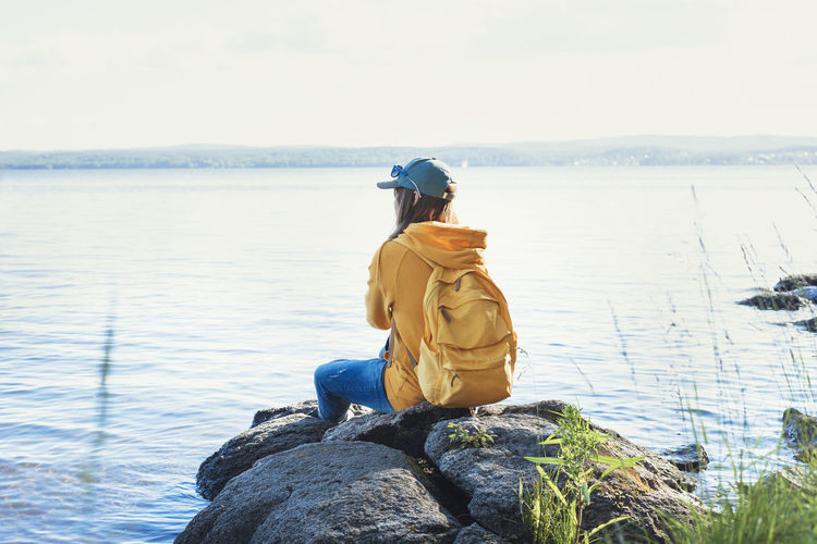 Young woman yellow hoodie with backpack sitting on rocks and looking at view of lake or river nature