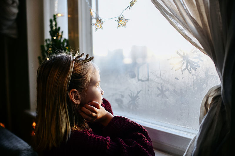 Side view of girl looking through window at home in christmas time