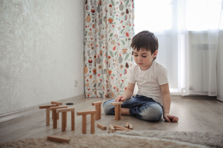 Boy caucasian brunette sad and serious plays on the floor, builds towers from wooden designer, 