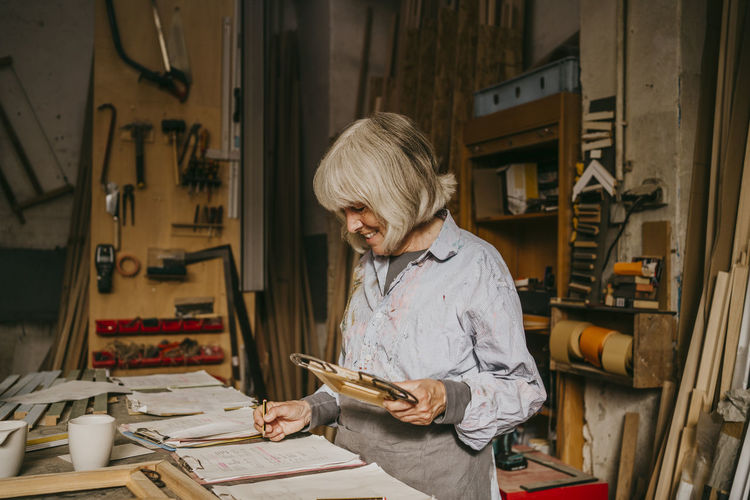 Smiling senior female carpenter holding frame while writing on clipboard at workshop