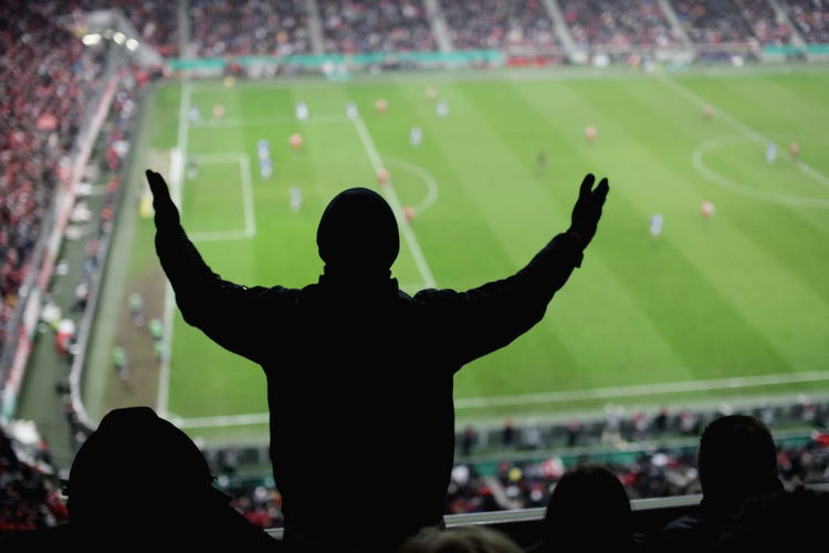 Rear view of spectator with arms outstretched standing at stadium during soccer match