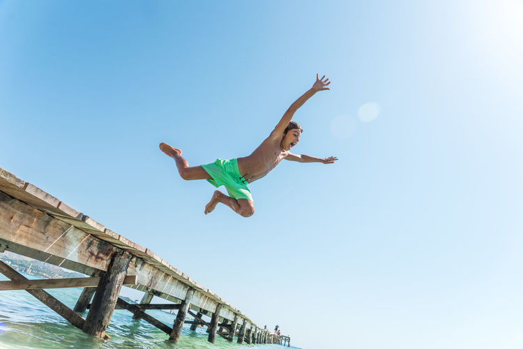 From below of cheerful teenage boy in moment of jumping from pier in sea enjoying summer weekend on sunny day