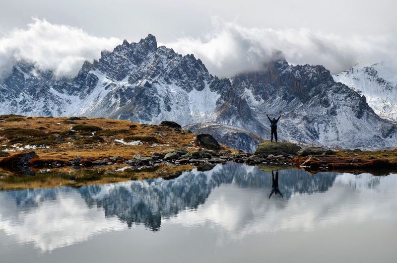 Man with arms raised reflecting on lake by mountains against cloudy sky
