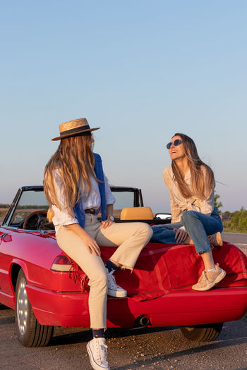 Happy female friends cheering sitting in a convertible red car at sunset time with golden light 