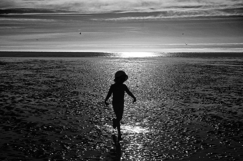 Full length rear view of silhouette child running at beach against sky