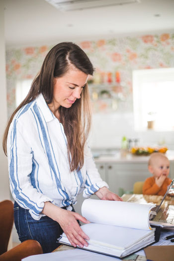 Busy female entrepreneur examining filed documents while daughter sitting in background at home office