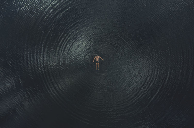 A man swims on his back in dark water. view from above. human provokes circular waves on the water. 