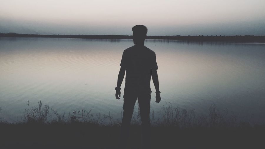 Rear view of silhouette young man standing by lake against sky during sunset