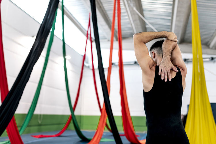 Back view of anonymous man in leotard bending and stretching arm while standing near colorful aerial silks during rehearsal