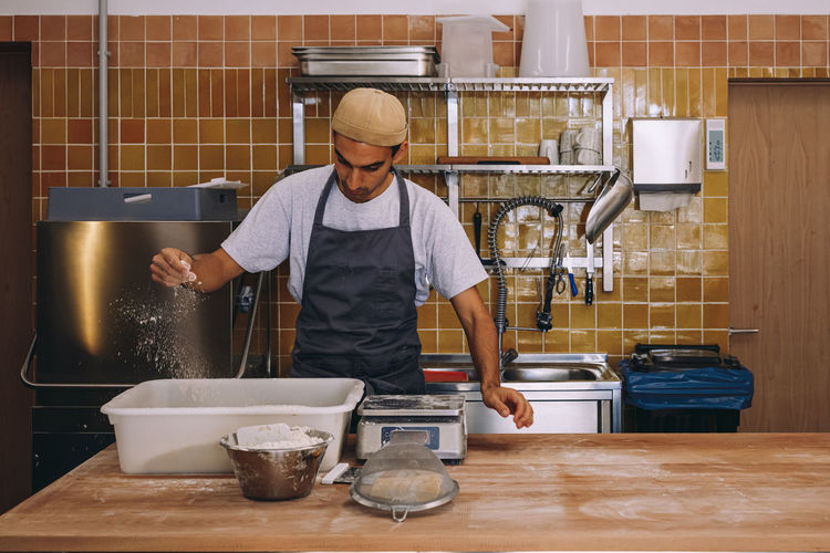 Positive young ethnic male baker in apron and hat sprinkling four on dough in big container while preparing bread in bakehouse