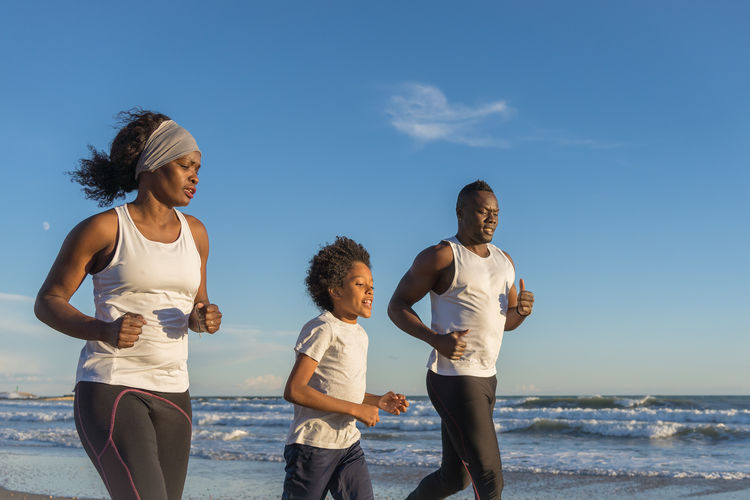 African american family in sportswear running along sandy beach near sea and training together at sunset in summer