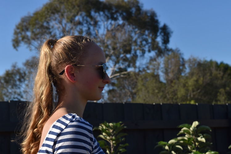 Side view of young woman in sunglasses standing at yard during sunny day