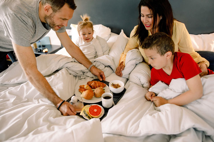 Father serving breakfast for family on bed in hotel room